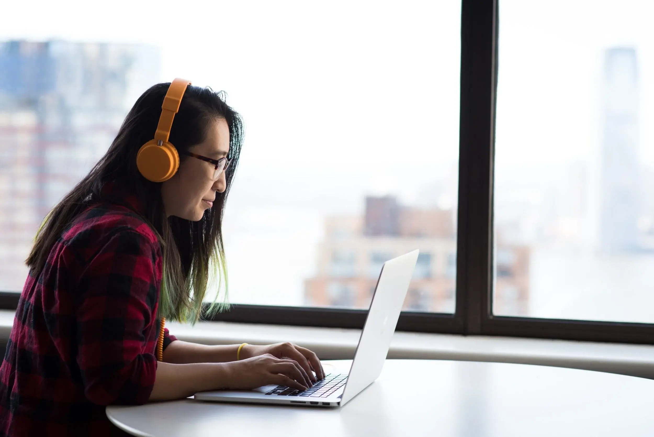A woman in orange headphones types on a laptop at a window overlooking a city skyline.