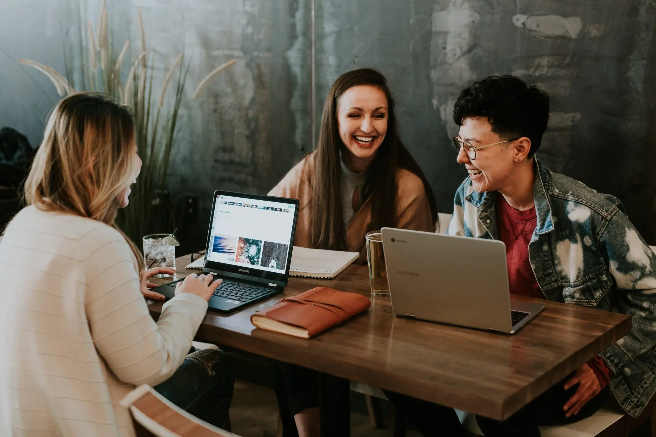 Three colleagues laugh together around a table with open laptops in a cafe.