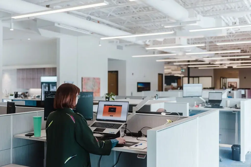 A woman works at a standing desk with a laptop and monitor in a bright open-plan office.