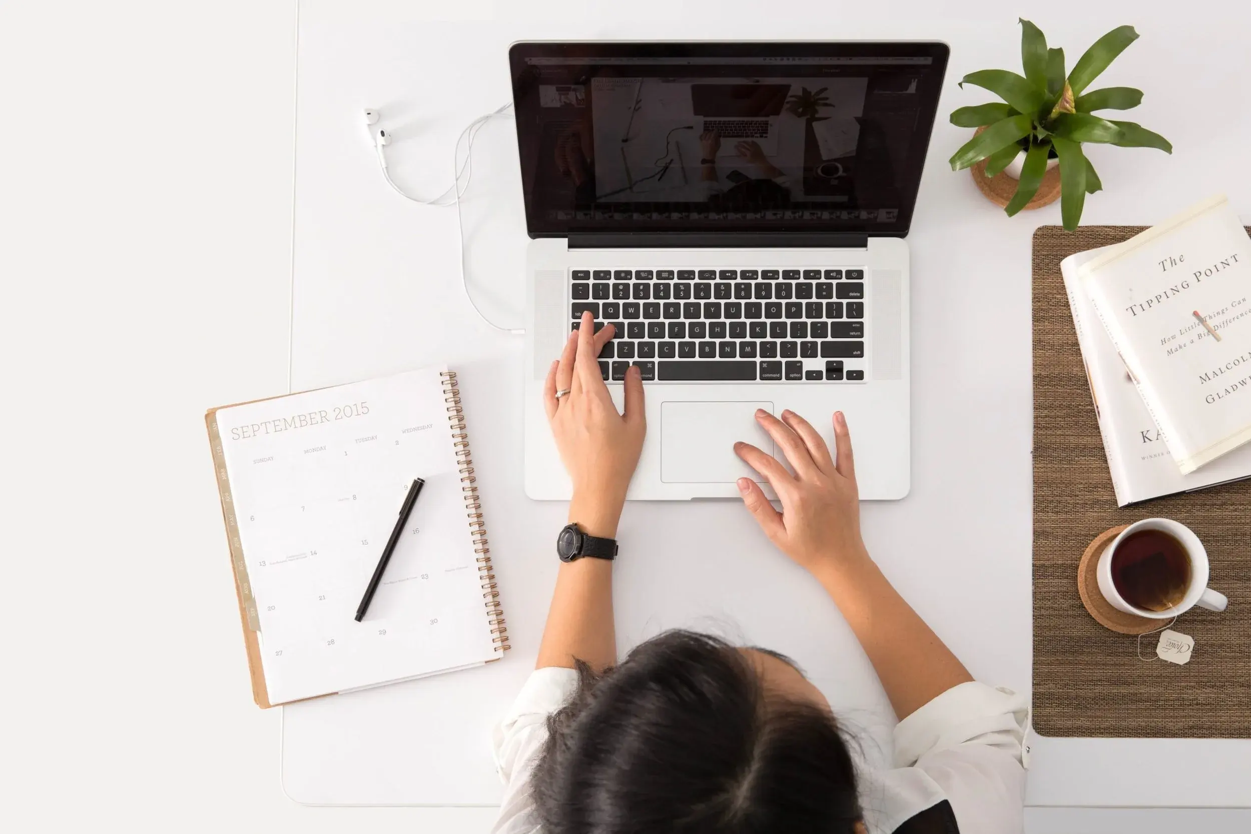 Overhead view of a desk: hands on a laptop beside a paper calendar, a pen, a plant and a cup of tea.