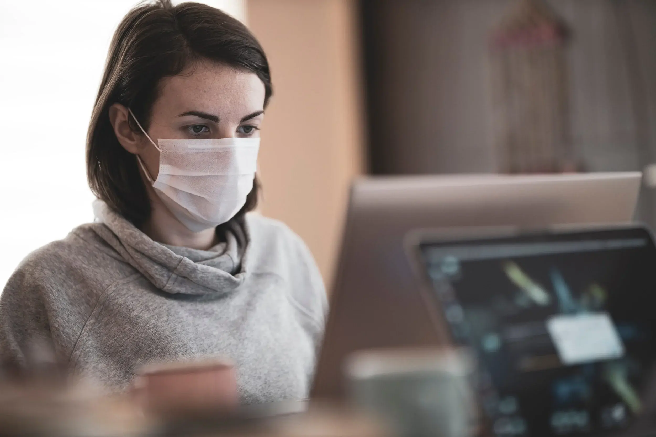 A woman wearing a surgical mask works at a laptop in a home office.