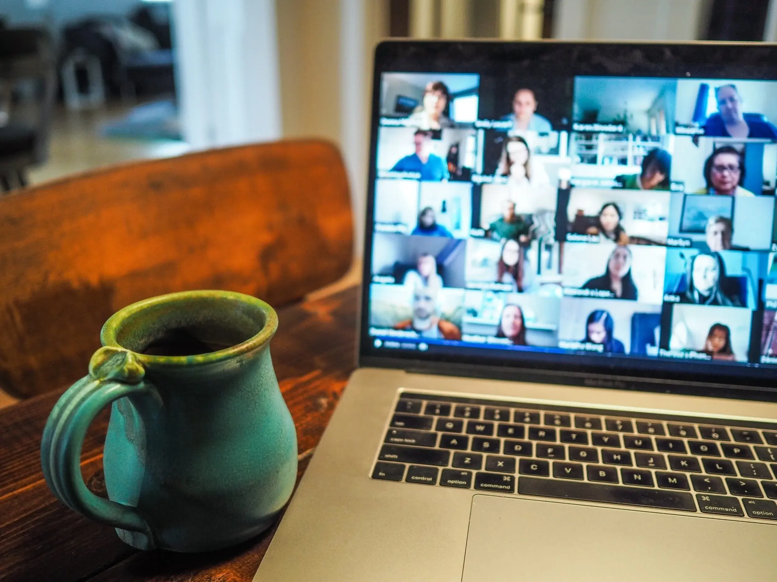 A laptop on a wooden table shows a grid of faces on a video call, next to a green ceramic mug.