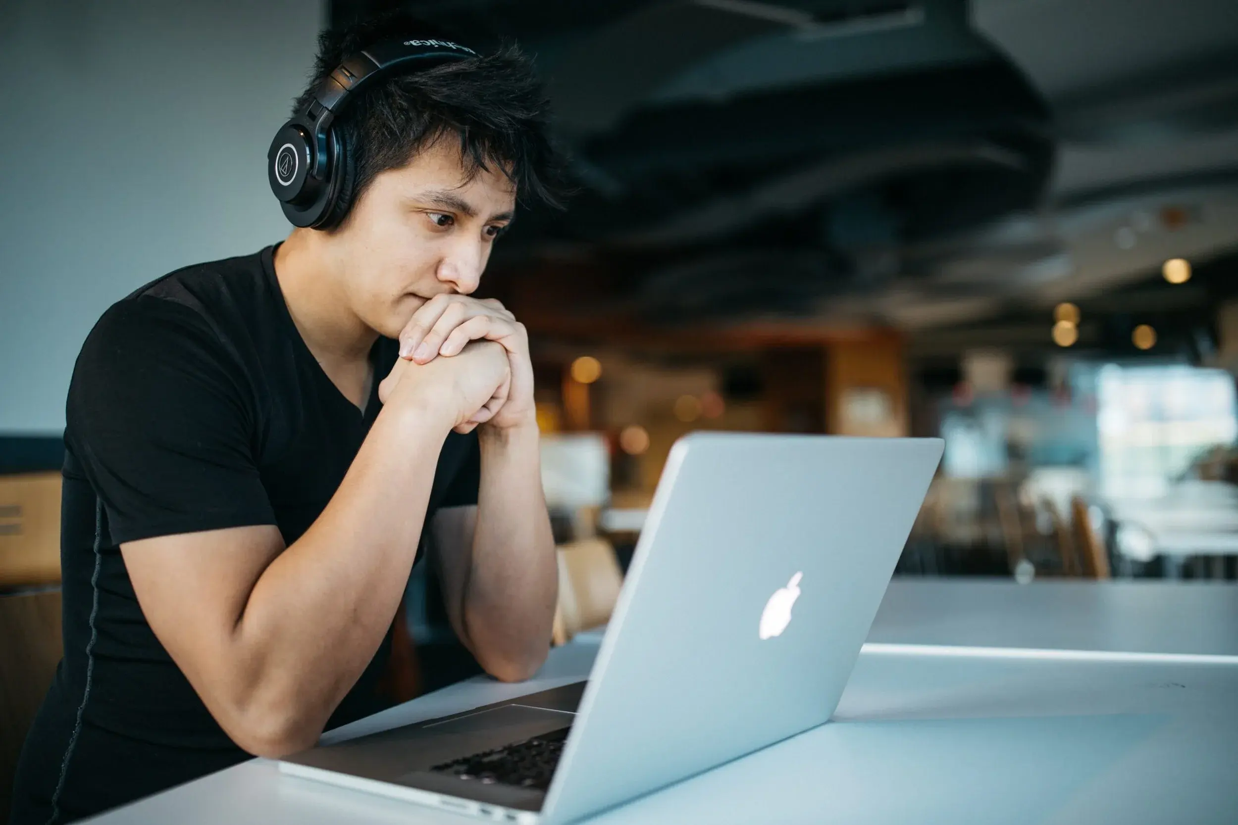 A student in headphones rests his chin on clasped hands, concentrating on a laptop screen in a cafe.