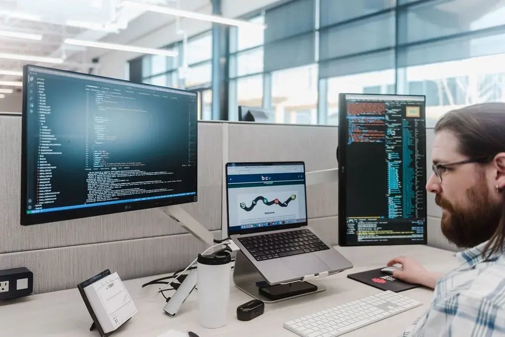 A developer at a desk with code on two monitors and the Business Career College course open on his laptop.