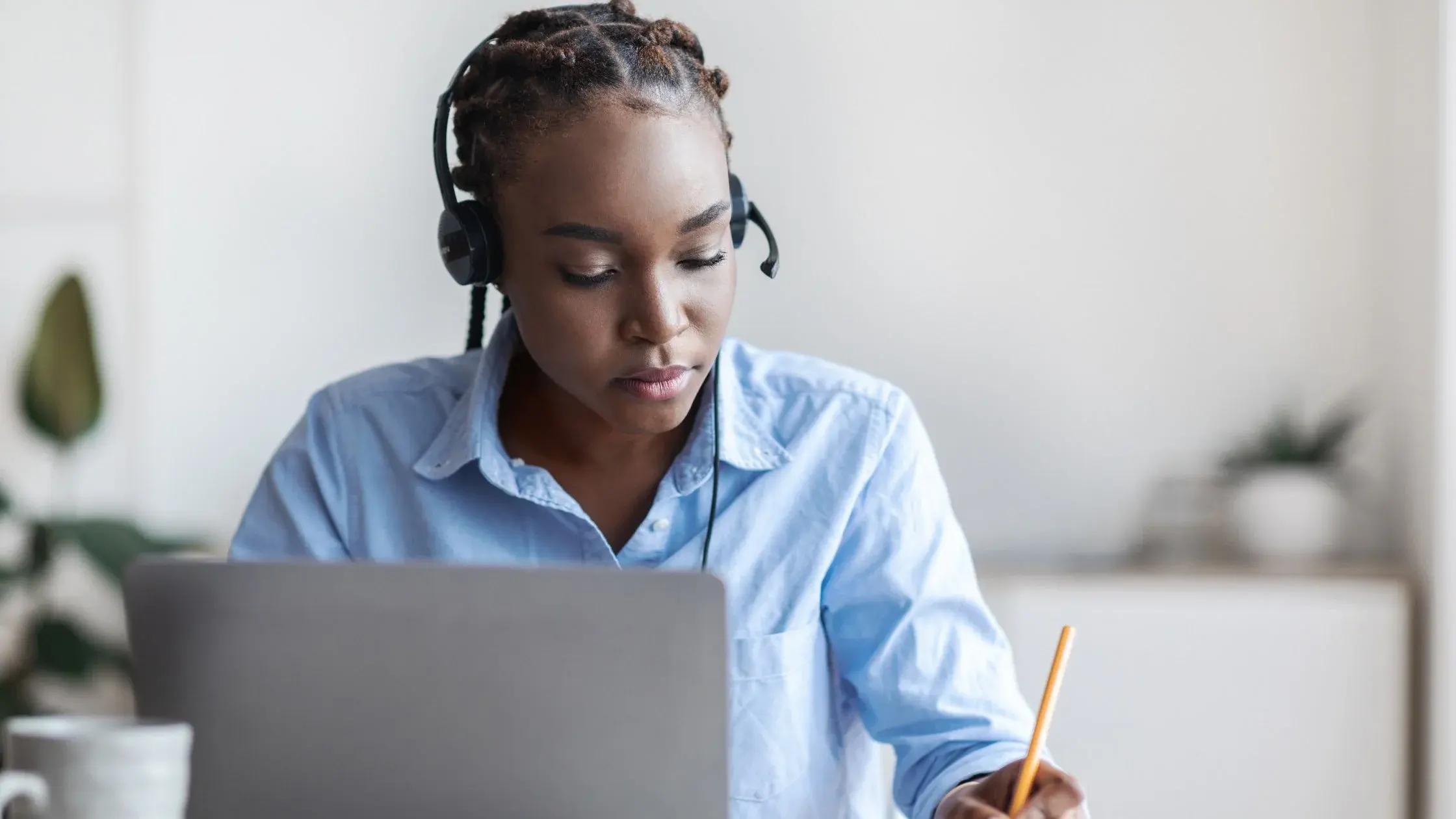 A woman wearing a headset takes notes with a pencil beside her laptop.