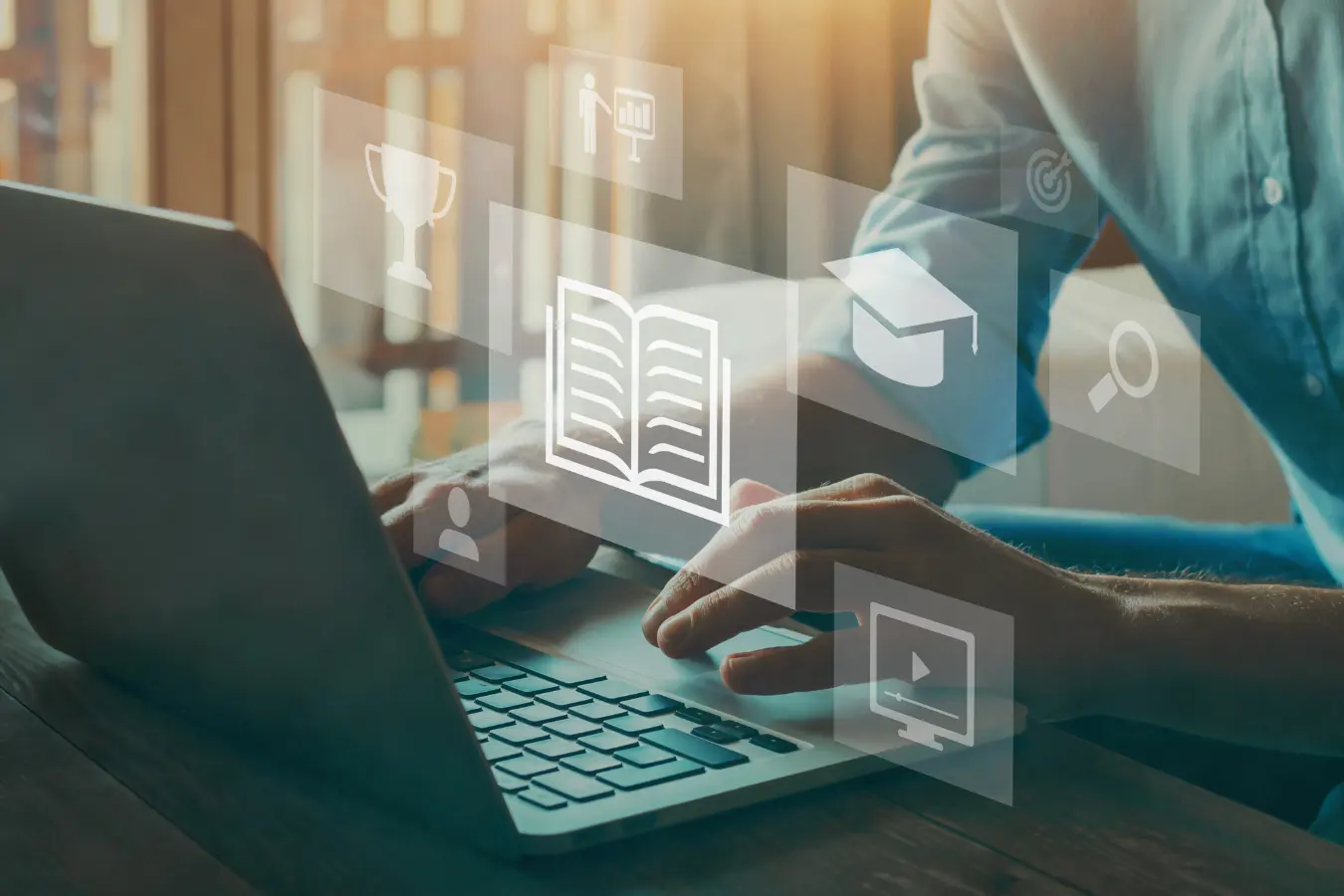 Hands typing on a laptop overlaid with e-learning icons: an open book, graduation cap, trophy and video player.
