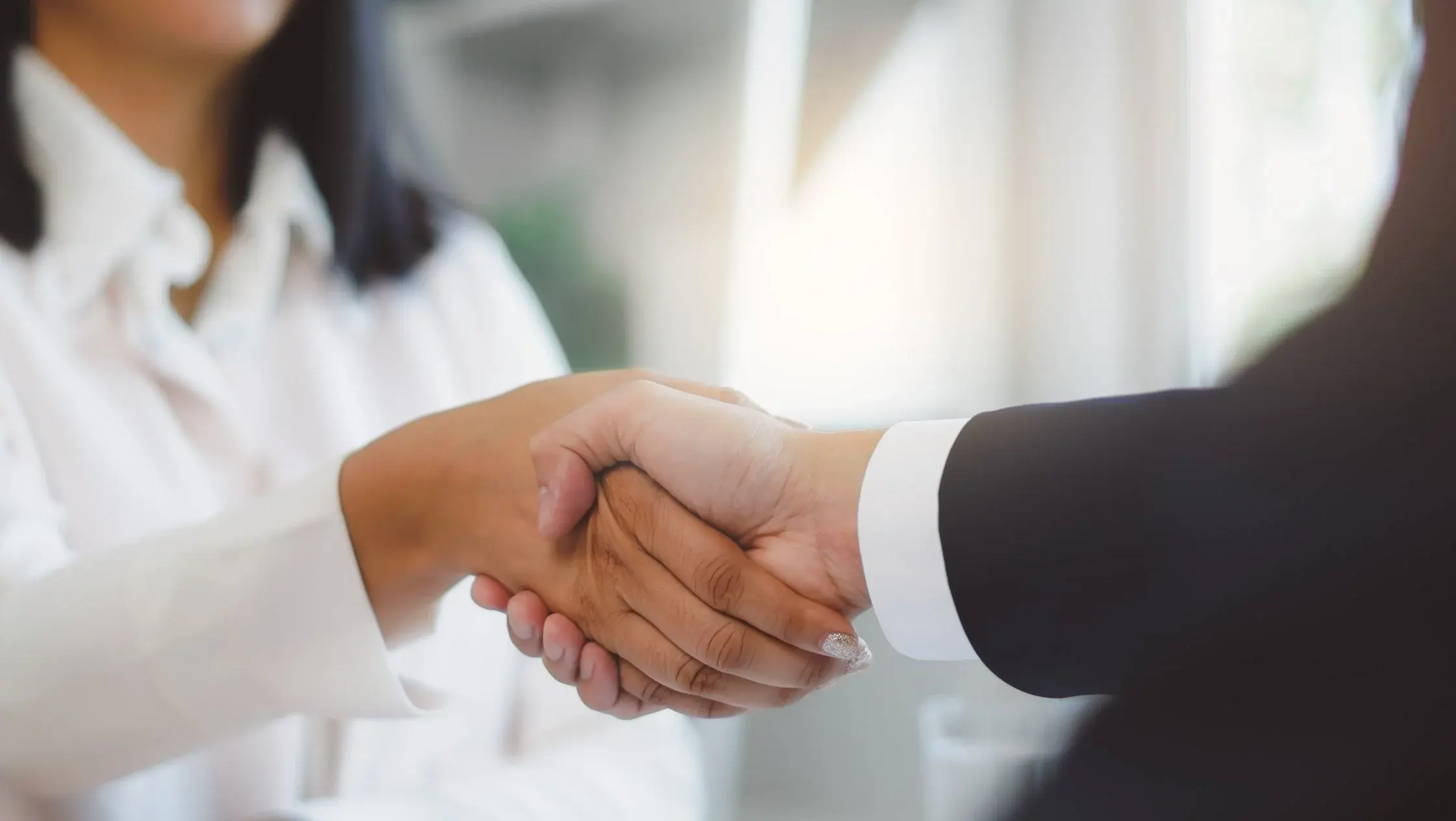 Two people in business clothing shake hands in a bright office.