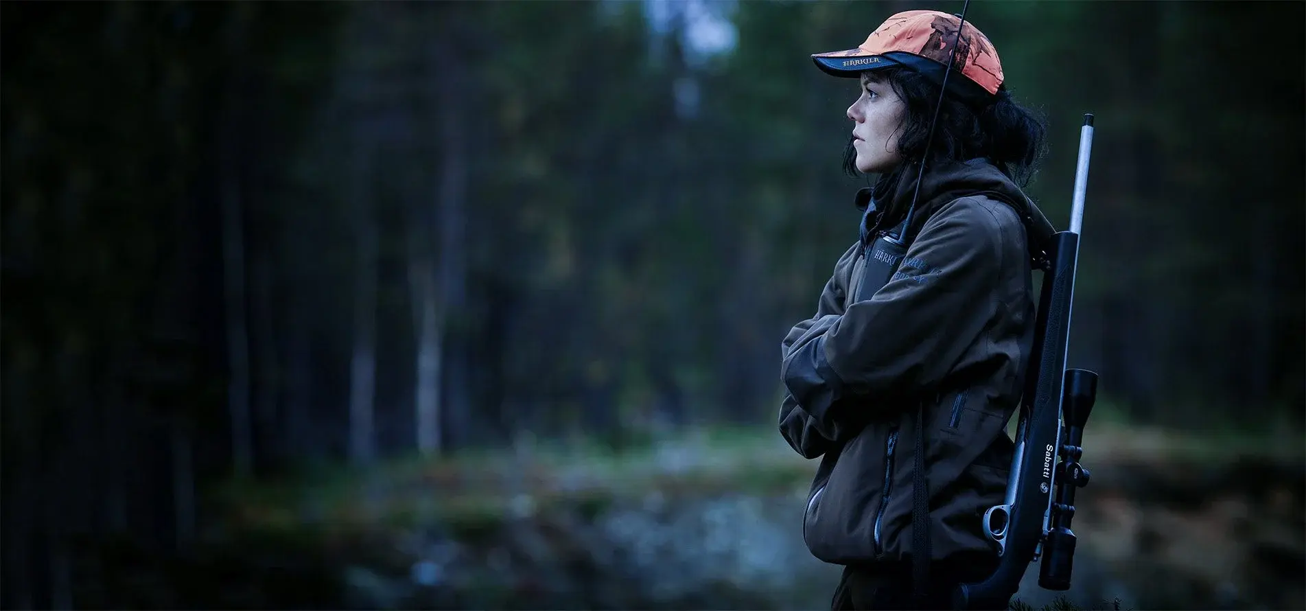 A hunter in an orange camo cap stands in a forest at dusk with a scoped rifle slung over her shoulder.