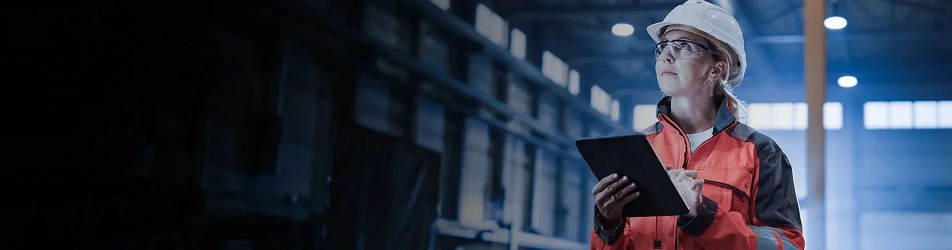 A worker in a hard hat, safety glasses and orange hi-vis jacket checks a tablet inside an industrial plant.