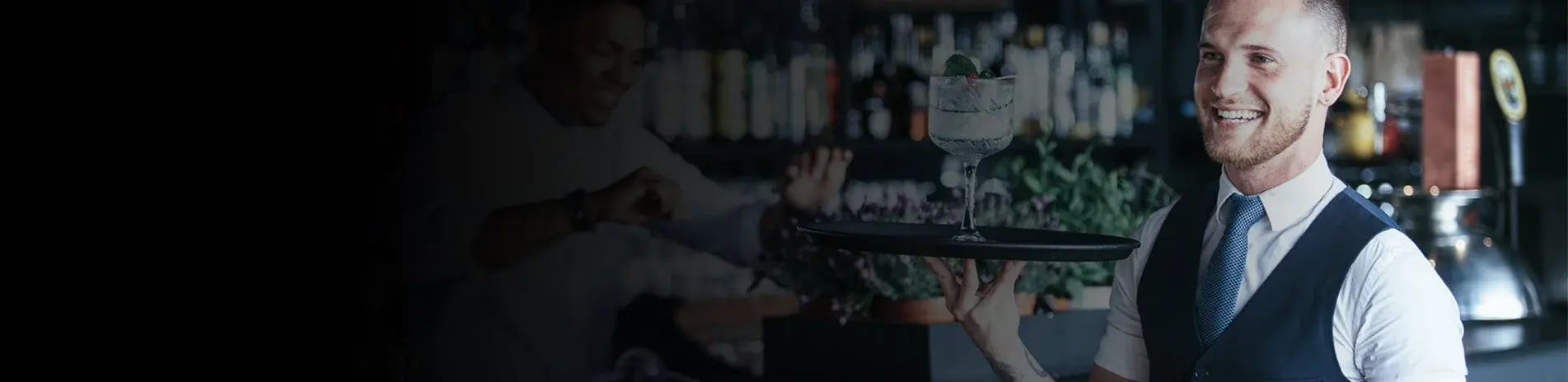 A smiling server in a waistcoat and tie carries a tray with a garnished cocktail past a stocked bar.